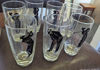 Front-angle photo showing seven clear drinking glasses with black silhouette golfing men in various poses, arranged in a hexagonal formation on a brown surface.