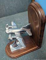 Single silvered metal airplane mounted on a metal stand attached to a curved wooden bookend base shown from front left angle.