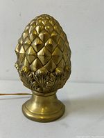 Front view of solid brass lamp shaped like a pineapple cone or artichoke with detailed scale texture and leaf design at the base on a round pedestal.