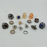 Full view of all 16 rings arranged on white background showing variety of styles, materials, and colors.