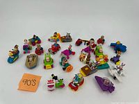 Wide view of 25 assorted colorful vintage toy figures and vehicles from the 1990s, placed on white background with a note indicating '90's'.