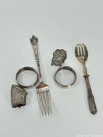 Six silver items arranged on a white background: two napkin rings, two forks, a leaf-shaped brooch, and a curved clip.