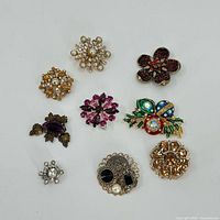 Full group of nine vintage brooches arranged on white background, showing variety of colors and styles including floral and jeweled designs.
