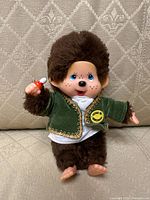 Front view of Monchhichi doll sitting on a patterned fabric surface, holding a baby bottle, showing green velvet jacket with gold trim and yellow patch.