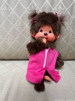 Front view of Monchhichi doll showing face, pink shirt partially open, thumb in mouth, and pink bows in hair.