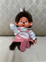 Front view of Monchhichi doll sitting on a patterned cushioned surface, showing facial details, outfit, and shoes.