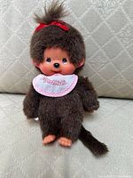 Front view of the Monchhichi doll showing brown fur, plastic face with painted eyes and nose, bib with text, and red bow on head.