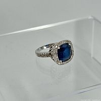 Image showing side angle of silver ring with cushion-cut dyed sapphire surrounded by clear stones and stones on band.