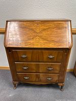 Front view of wooden secretary desk with closed fold-down top showing symmetrical wood grain pattern and three drawers with metal handles.