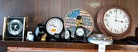 Photo of the 10 clocks arranged on a shelf, showing a variety of analog and digital clocks including a large round wooden framed wall clock, a decorative Americana themed round wall clock, and several smaller desk clocks.