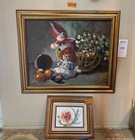 Both framed oil paintings as displayed on wall, larger painting with Raggedy Ann dolls and basket of flowers, smaller floral painting below it.