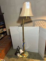 Full view of tall brass floor lamp with beige fabric shade, showing the lamp standing on tile floor near brick wall.