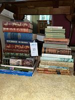 Wide shot showing stacks of varied vintage and mid-century books in different bindings including leatherbound and hardcover, arranged under a wooden table.