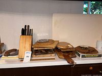 Photo of wooden knife block with multiple knives, a small wooden cutting board, and wooden hotplates on a countertop