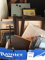 Box filled with assorted picture frames made of wood, metal, and plastic. Frames vary in size including large, medium and small sizes.