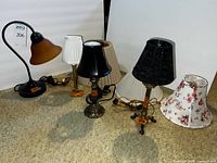 Four table lamps and three additional lamp shades shown on carpet. Includes the black metal desk lamp with amber mottled glass shade, and three traditional styled lamps with varied shades including pleated cream, black fabric, and floral fabric.