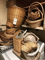 Woven baskets stacked showing various sizes and materials including wicker, rattan, bamboo, and reed. Large round basket with attached tag visible and smaller baskets nested inside and around it.