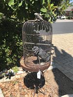 Full view of rusty round metal bird cage on metal stand with shadow cast on ground in outdoor setting