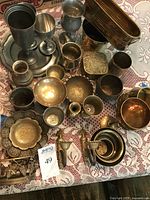 Photo showing variety of brass, copper, and pewter goblets, bowls, plates, candle sconces, and trays arranged on lace tablecloth.