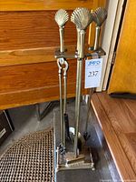 Full view of brass fireplace tool set with shell motif handles and matching stand on hardwood floor next to wooden furniture.