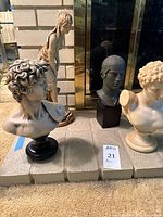 Four classical-style sculptures, three busts and one full figurine, displayed grouped together on a floor with carpet and brick wall background.