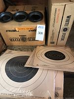 Two boxes of White Flyer ceramic clay pigeons sitting on a shelf with several paper shooting targets laid out in front.