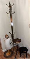 Tall brass coat rack with multiple hooks, small dress form torso with decorative wreath and lace, small hexagonal wooden table, small black wooden stool grouped in corner