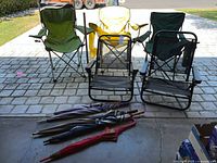 Five collapsible camp chairs in various colors and five large closed umbrellas laid in front of them.