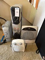 Four electric heaters grouped on carpet floor including Costway oil radiator, ProFusion space heater, Pelonis fan heater, and Living Solutions smaller tower heater.
