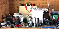 Wide photo showing assortment of emergency radios, lantern, flashlights, tote bag, and other tools on wooden surface with pegboard background.