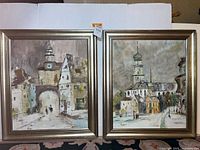 Set of two framed cityscape paintings showing town scenes with clock towers and architecture, silver-toned frames, signed by artist Riggio.