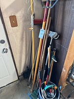 Photo showing assorted long-handled garden and yard tools including rakes, shovels, hoes, broom, and squeegee leaning against a wall near a door, with a bucket on the floor containing various hand tools.