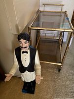 Front angle of gold finish metal bar cart with two glass shelves next to waiter statue figurine
