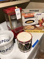 Red and clear popcorn maker with Home Pride branding, included instruction manual, set next to two white popcorn bowls and toy drum ice bucket. Also visible is Nostalgia Electrics party maker oven box with illustrated hot dog and baked goods.