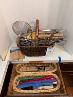 Wicker basket filled with various kitchen utensils including wooden handled knives, wire skimmer, metal tongs, placed on a shelf alongside two small wicker baskets containing colorful knives.