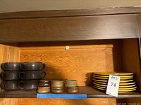 Stack of six brown handled stoneware soup bowls, six small stoneware dishes in two earth tones, and six yellow stoneware plates stacked apart on a wooden shelf.