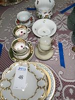 Overview of assorted teacups, saucers, plates, and a porcelain bucket showing varied patterns and gold accents on a lace tablecloth.
