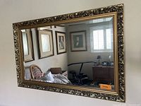 Rectangular wall mirror with ornate gold-toned carved frame, reflecting a room interior. The frame features detailed Baroque or Victorian styling with floral and scroll elements.