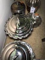 Photo showing two covered silver plated chafing dishes with ornate, scalloped rims and decorative handles, plus a pedestal basket with intricate openwork design at the back.