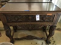 Front view of the intricately carved wooden side table with ornate detailing on drawer front, cabriole legs and cross stretcher base visible.