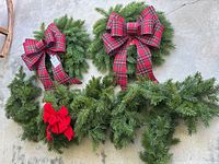 Two 18-inch faux evergreen wreaths, one with a red plaid bow and one with a smaller red fabric bow, and several sprigs of faux evergreen swag arranged on concrete.