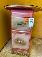 Front view of a small painted 2-drawer cabinet with pink and gold distressed colors, gold handles, decorative carvings on base, and a round glass top.