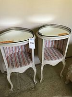 Pair of vintage white painted wood side tables with scalloped legs and bottom, oval mirrored tops with loose removable mirrors and decorative metal rims. Each has an open cubby lined with floral fabric in stripes of pink and beige.