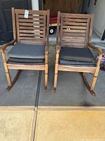 Pair of brown finished wooden rocking chairs with gray cushions, shown outdoors on concrete. Slat-back style and visible wear on wood.