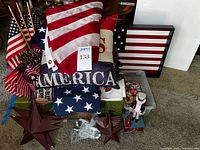 Overview photo showing stacked patriotic pillows, two American flags, rust-look stars, framed flag artwork, and some figurines