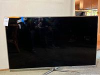 Front view of TV on floor with attached remote at top and silver V-shaped stand visible