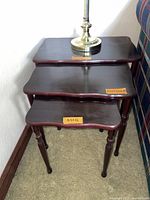 View of 3 cherry wood dark finish nesting tables stacked and standing in a corner next to a sofa. Shows table tops and turned legs; scalloped edges visible. Size labels on tops.
