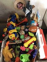 Top view of assorted vintage and modern children's toys packed in a box, showing wooden puppet, plastic Fisher-Price toys, plush toys and sports themed toys.