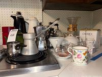 Photo showing various vintage kitchenware items including thermal carafes, moka pot, enamel and aluminum stove-top milk warmers, a tea pot, and Chemex coffee maker with filters on a kitchen counter.