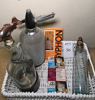 Overview of vintage seltzer bottle set with metal mesh covered siphon bottle, glass seltzer bottle with fish stopper, plain glass bottle, and multiple boxes of carbonation chargers.